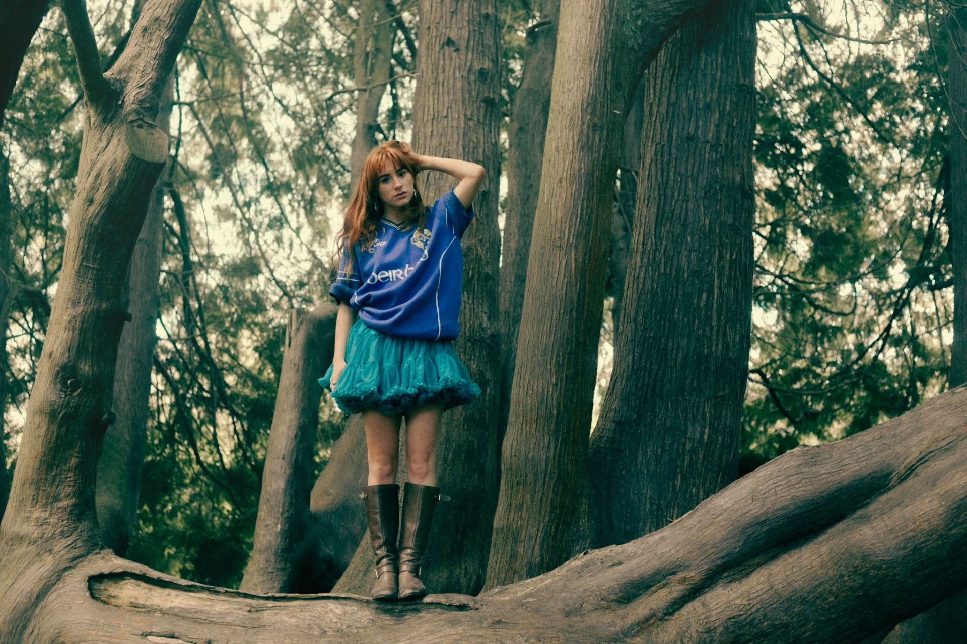 Aisling Sharkey standing on a fallen tree in the woods, wearing a skirt and football shirt