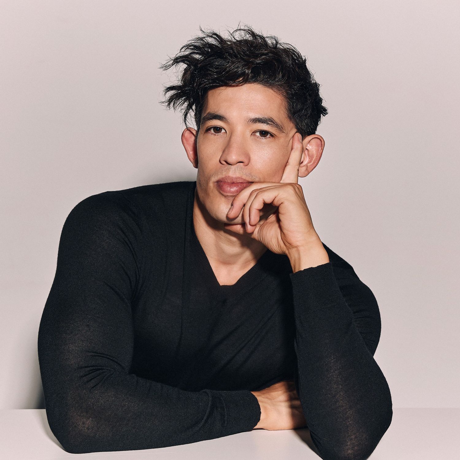 Australian actor Chris Chung in a tight black long sleeve t shirt, looking at the camera, resting his head in his hand
