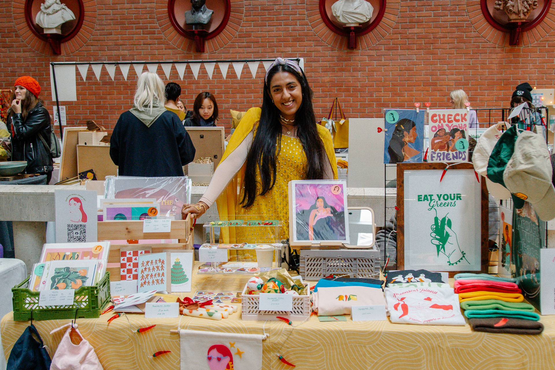 A young wman presenting a table of art prints, cards and t shirts