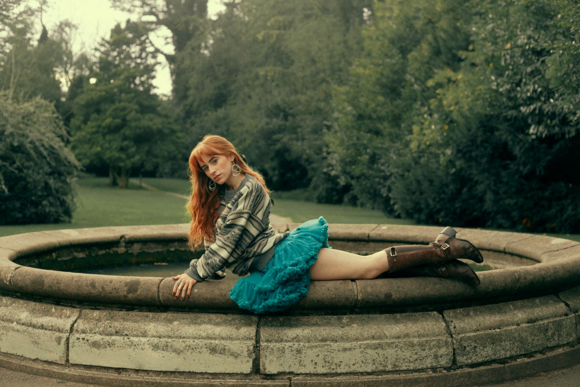 Aisling in a jumper, skirt and boots, lying on the side of a fountain