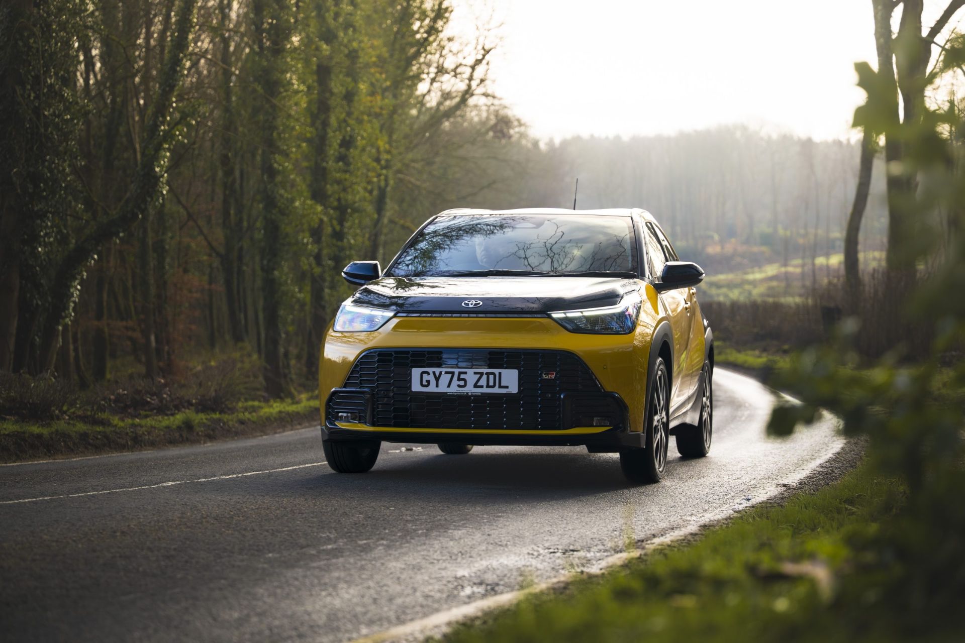A yellow Toyota Aygo X driving on a treelined road